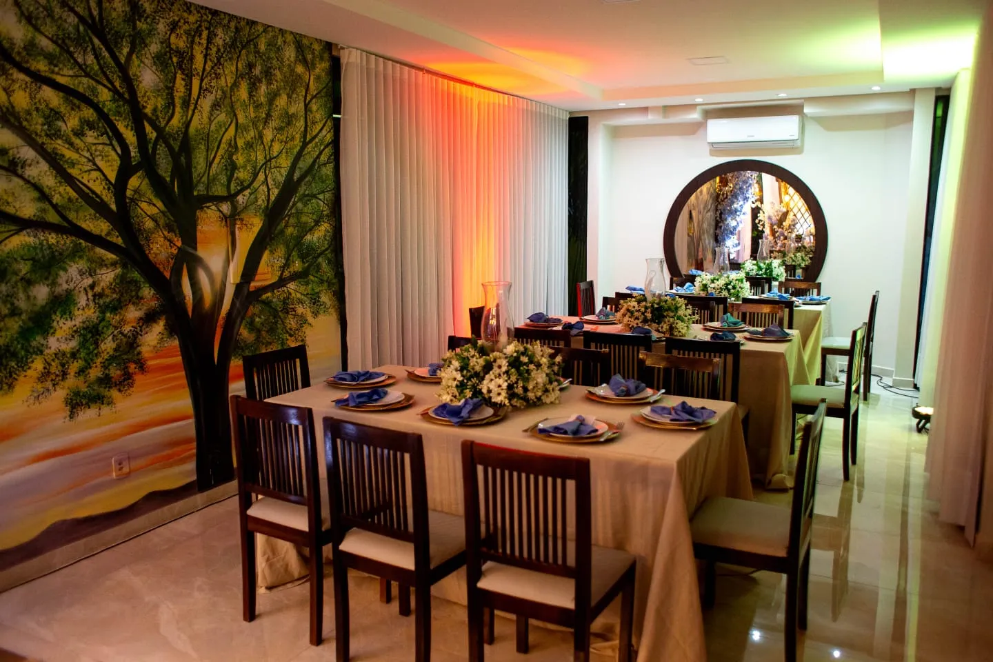 Salão de eventos com mural de seringueiras pintado à mão e mesas postas